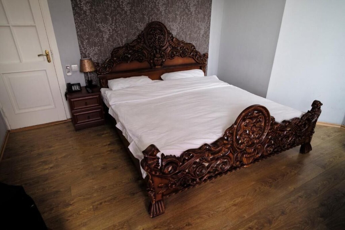 A wooden bed with intricate carvings, a bedside table with a lamp, and a patterned wall.