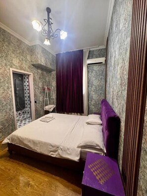 A bedroom with a bed, a purple headboard, a wooden floor, a chandelier, and a window with curtains. - Hotel Kalanga (T'bilisi)