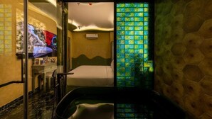 Quarto de hotel com cama, banheira envidraçada, toucador com espelho e parede com padrão geométrico.
