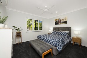 Una camera da letto con un letto, comodini, una ventola a soffitto, una finestra con persiane, una pianta e un quadro appeso al muro.