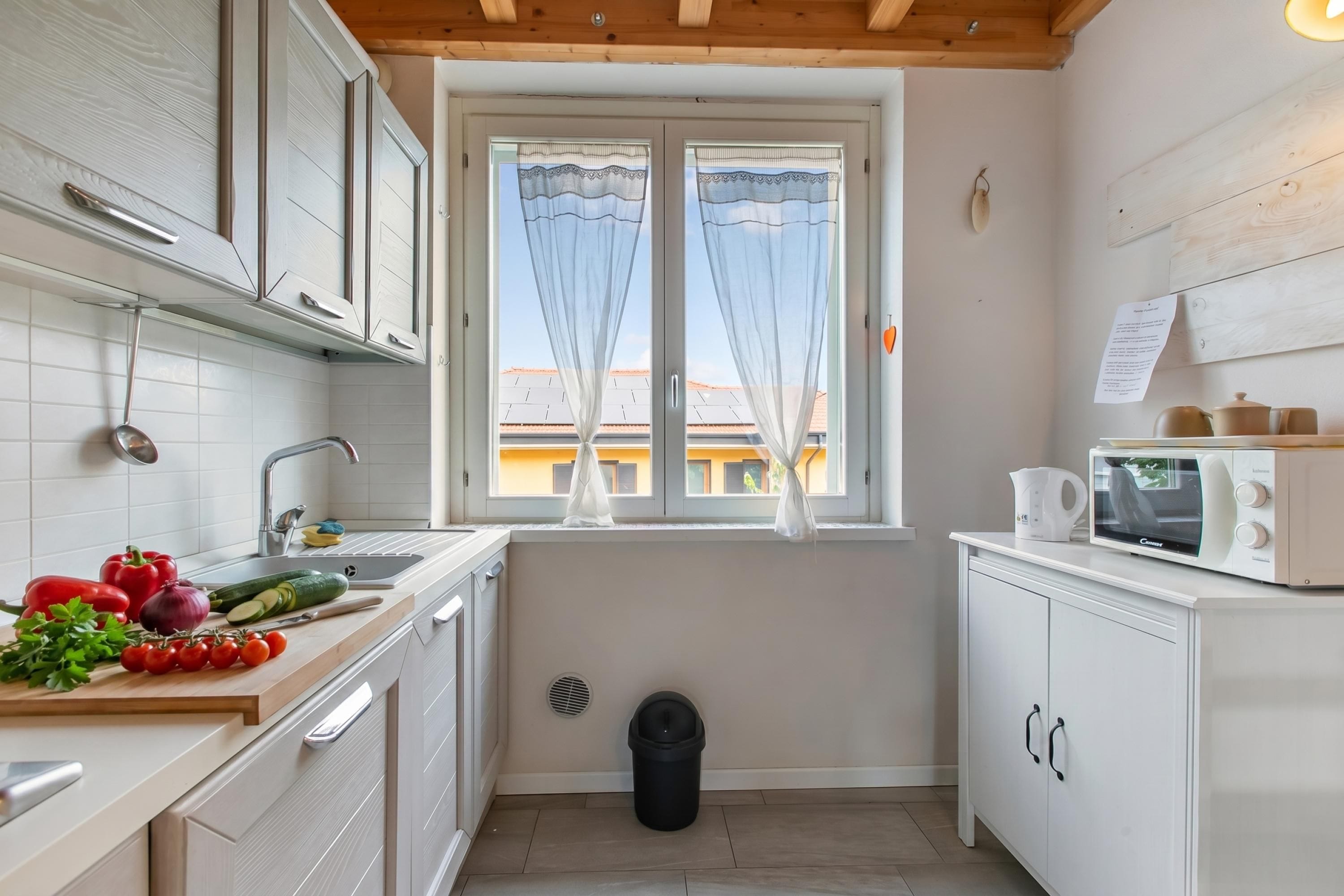 A compact kitchen with white cabinets, a window with sheer curtains, a microwave, and a sink.