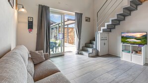 A modern living room with a sofa, a staircase, a TV, and a sliding glass door leading to an outdoor area. - Cozy Ispra Residence With Terrace, Near Beach Air-con (Ispra)