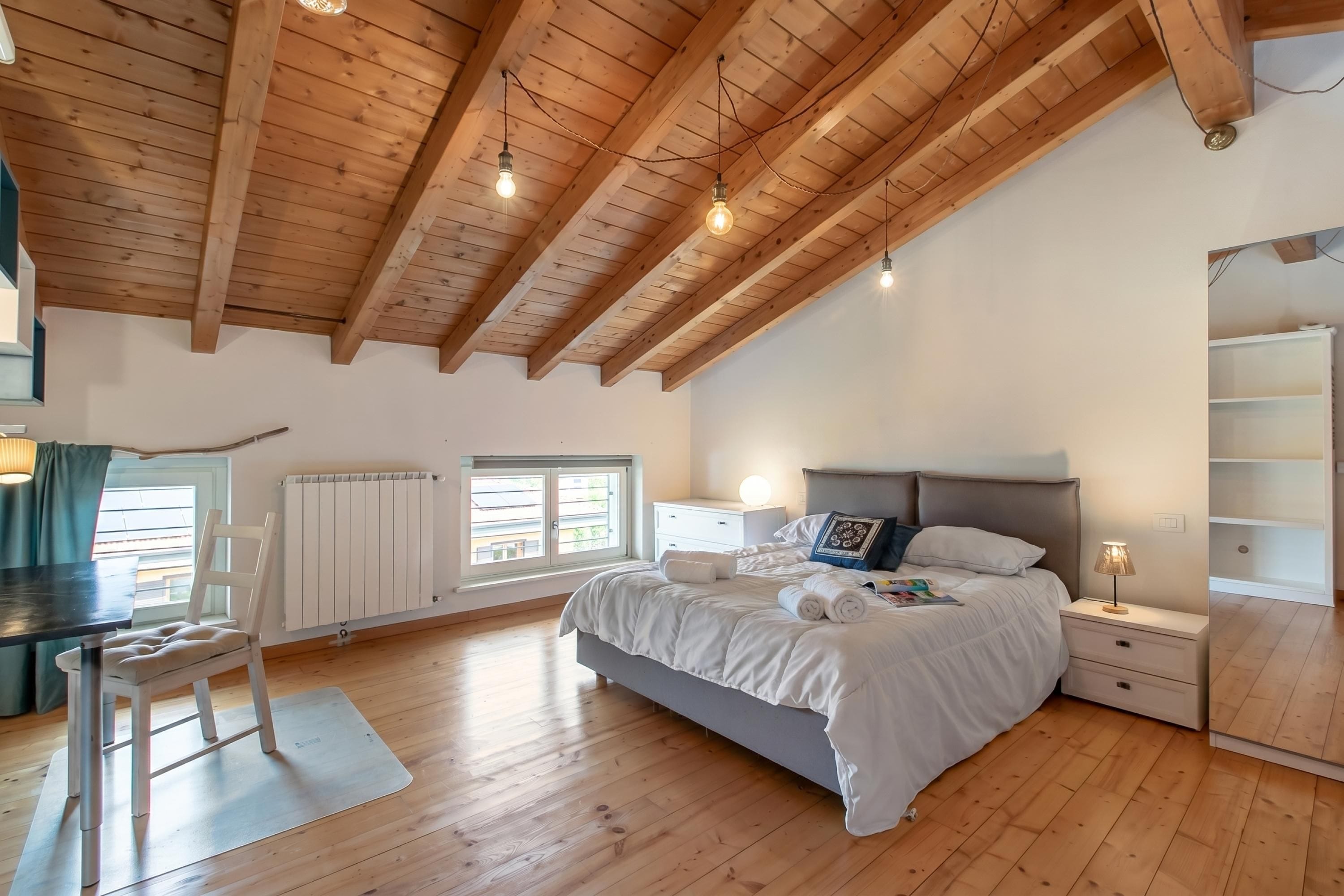 A bedroom with a wooden ceiling, a bed, bedside tables, a chair, and a radiator.
