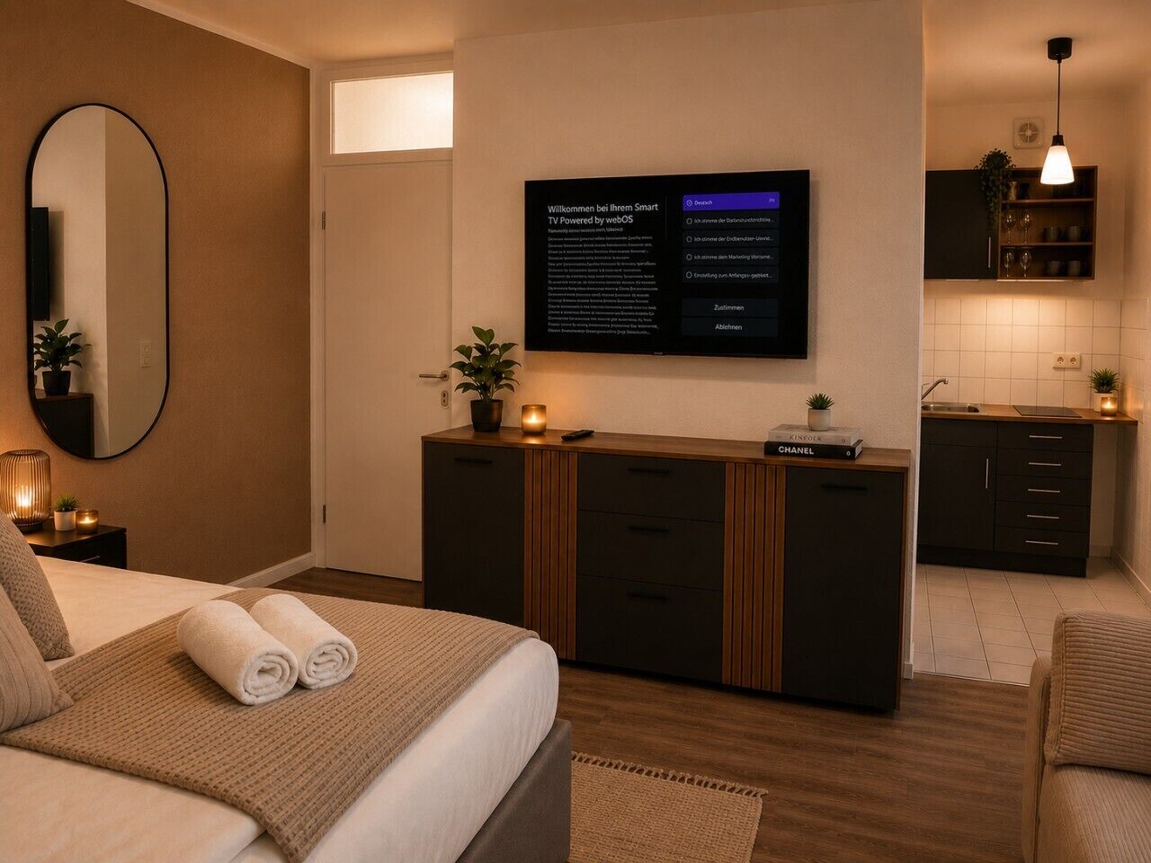 Ein modernes Hotelzimmer mit einem Bett, einem Nachttisch mit Lampe, einem an der Wand montierten Fernseher und einer kleinen Küchenzeile.