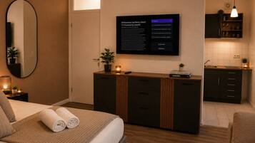 Ein modernes Hotelzimmer mit einem Bett, einem Nachttisch mit Lampe, einem an der Wand montierten Fernseher und einer kleinen Küchenzeile.