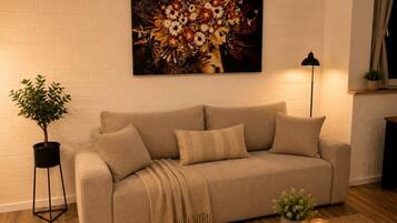Ein Wohnzimmer mit einem beigen Sofa, einer schwarzen Stehlampe, einem Topf mit Pflanze und einem Bild an der Wand.