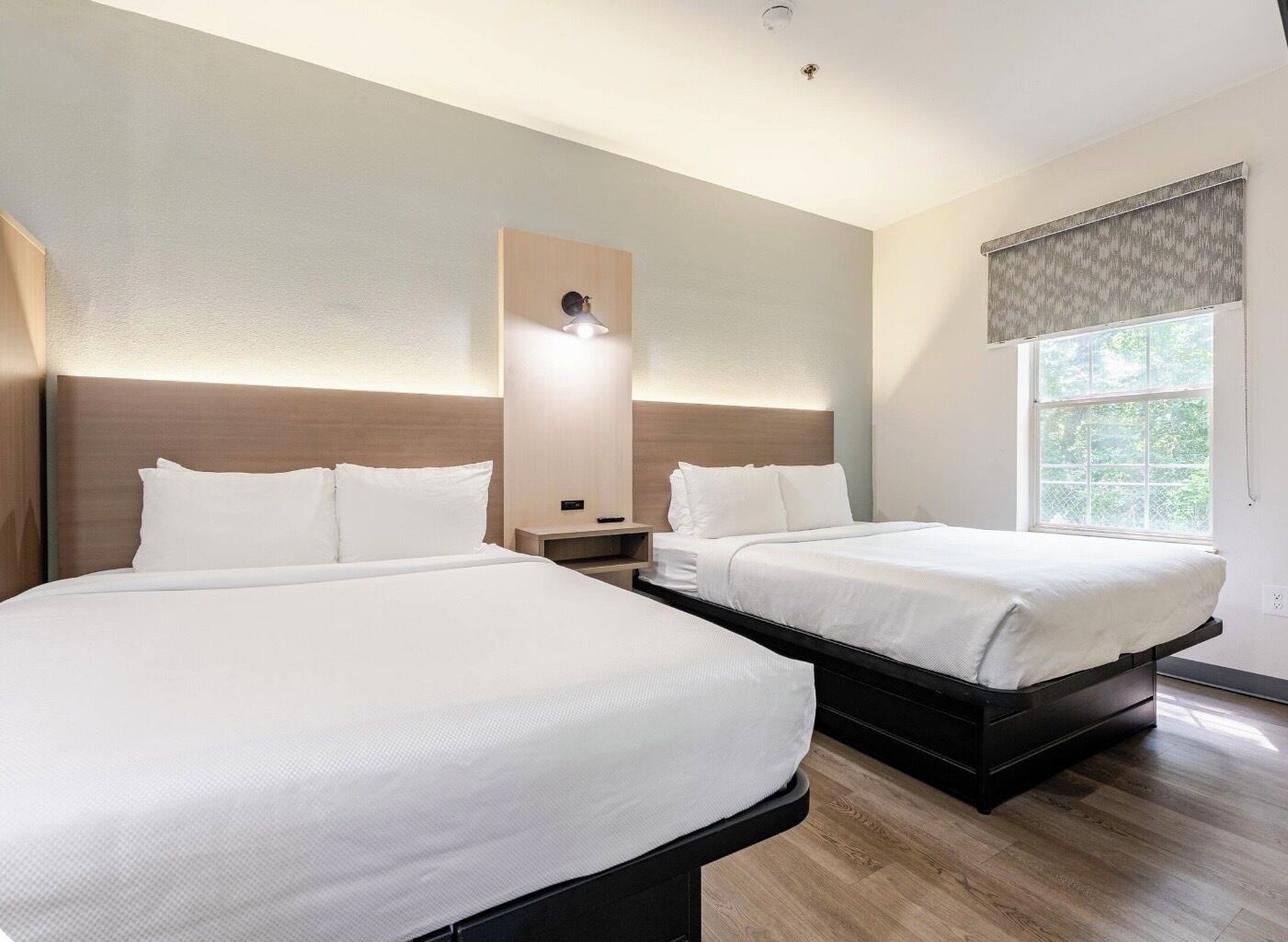 A hotel room with two beds, a nightstand, a wall-mounted lamp, and a window with a view.