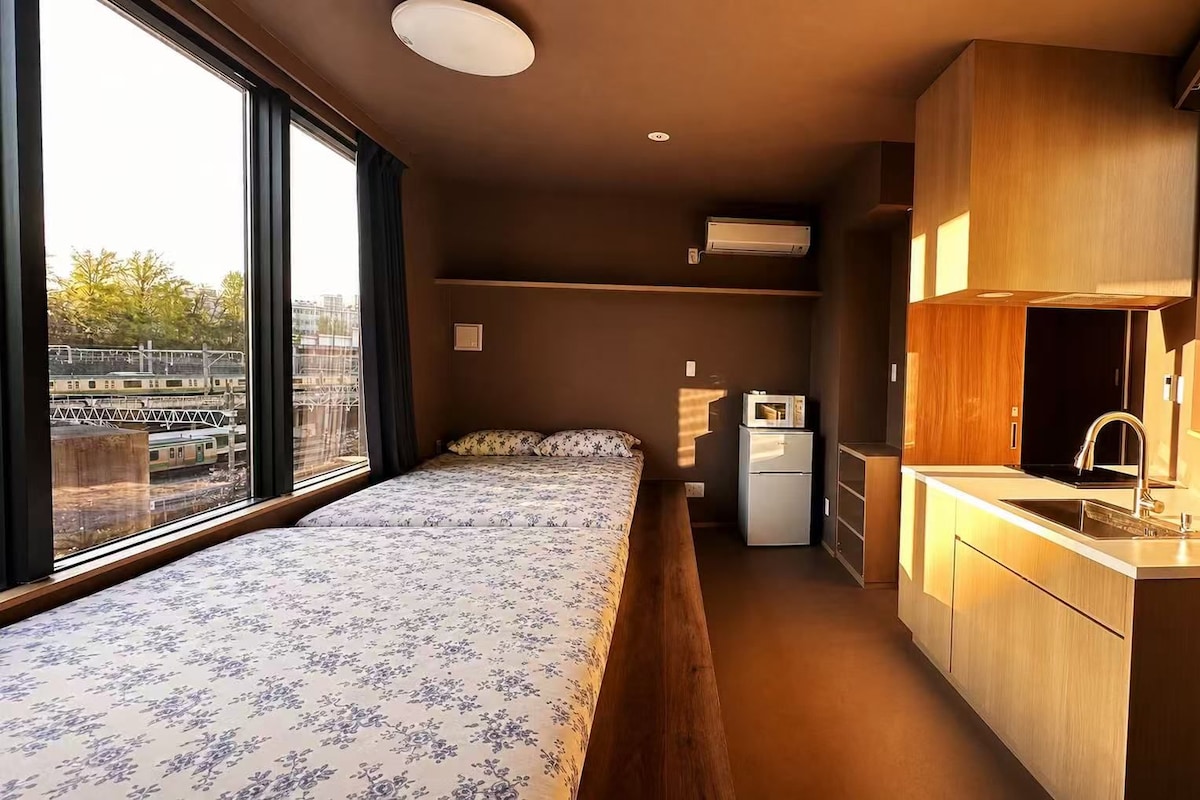 A compact room with a bed, a small kitchenette, and a window with a view of a train track.