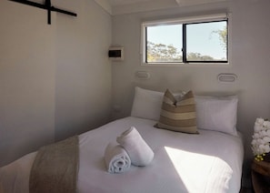 A neatly made bed with pillows, a rolled towel, and a window with a view of trees.