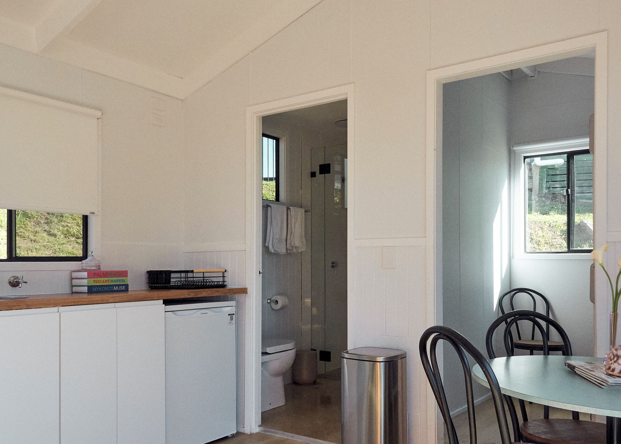 A compact kitchen with a dishwasher, a small refrigerator, a dining table with chairs, and a view of the outside through a window.