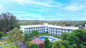 A multi-story hotel with a swimming pool, surrounded by lush greenery and a clear sky.
