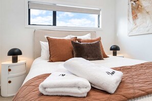 Una cama bien hecha con colcha marrón, almohadas blancas y una toalla blanca con un logotipo. Hay dos mesitas de noche con lámparas, y una ventana sobre la cama.