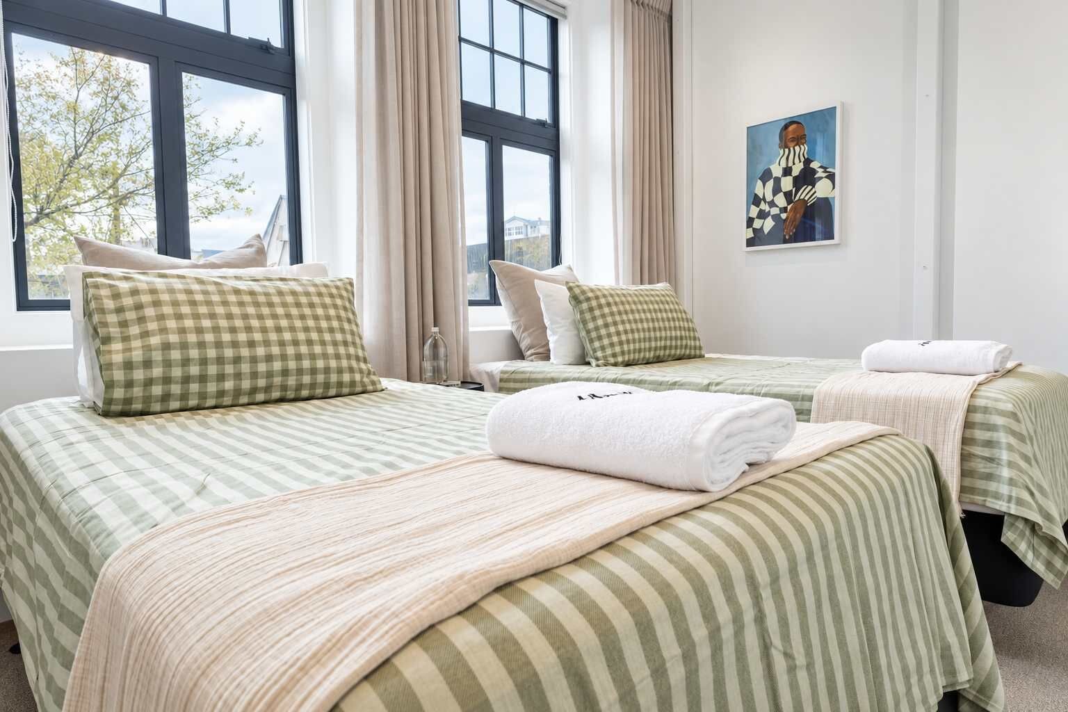 A hotel room with two beds, each with a striped bedspread and checkered pillows. There is a painting on the wall and a window with curtains.