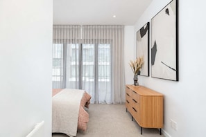 A modern bedroom with a bed, a wooden dresser, and two abstract paintings on the wall. - The Windsor | Wellington CBD | Stylish 3BR (Te Aro)