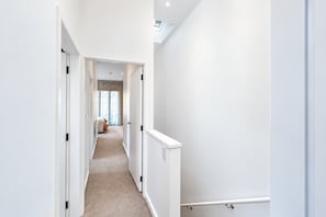 A narrow hallway with white walls and a carpeted floor leading to a bedroom with a bed and curtains.