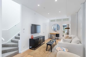 Ein modernes Wohnzimmer mit Treppe, Flachbildfernseher, rundem Spiegel, Couchtisch und weißem Sofa.