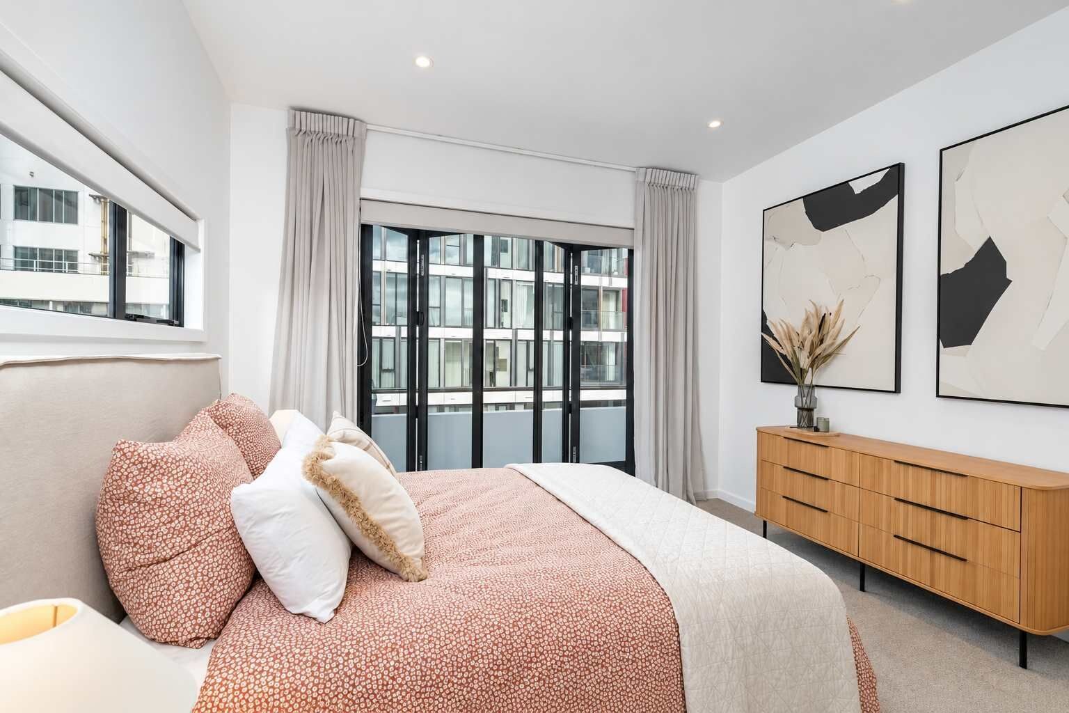 A modern bedroom with a large bed, a wooden dresser, and a city view through the window.