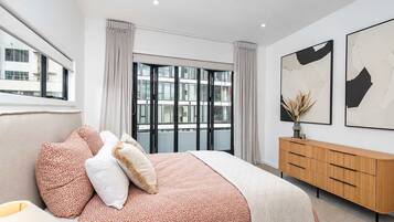 A modern bedroom with a large bed, a wooden dresser, and a city view through the window.