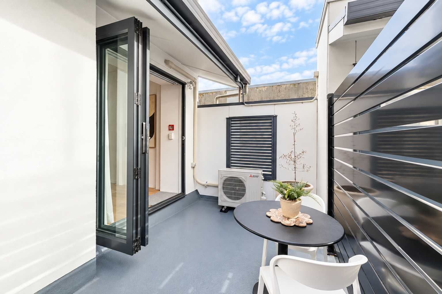 A small outdoor patio with a table and chairs, a potted plant, and a sliding glass door leading to an interior space.