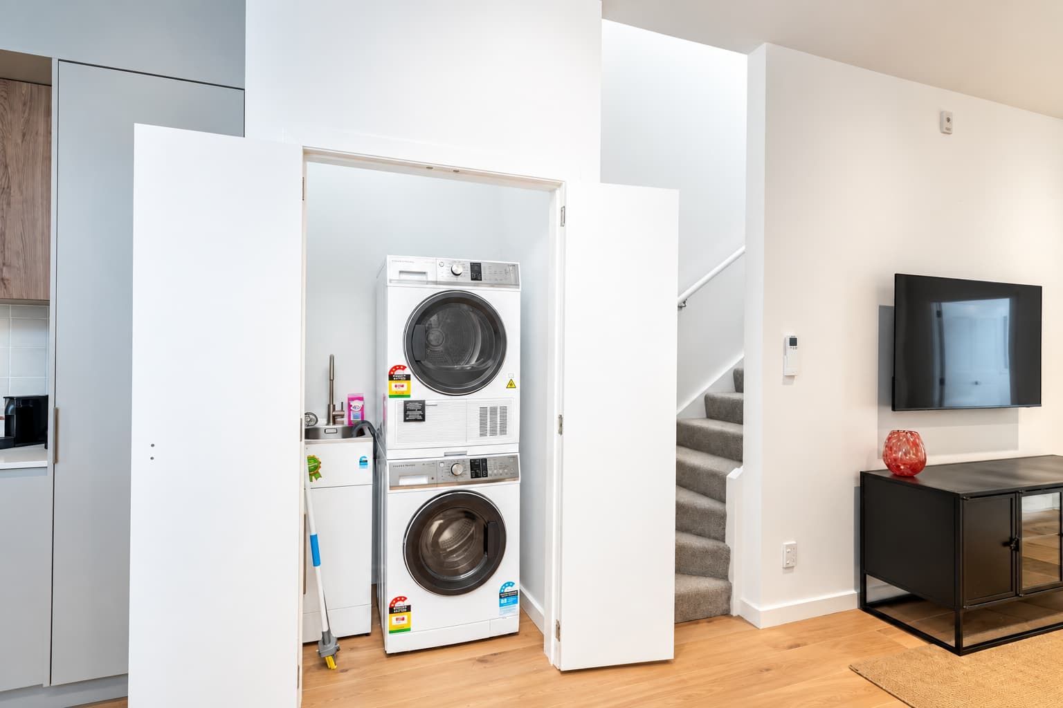 A laundry room with two stacked washing machines, a staircase, a TV mounted on the wall, and a small cabinet with a decorative item.