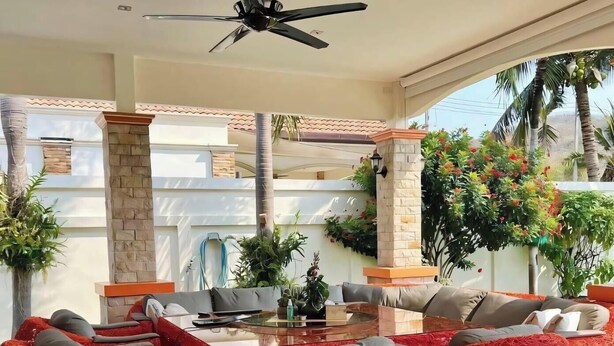 Zona esterna moderna con sedute in vimini rosso, ventilatore a soffitto e vista su una casa con tetto in tegole.