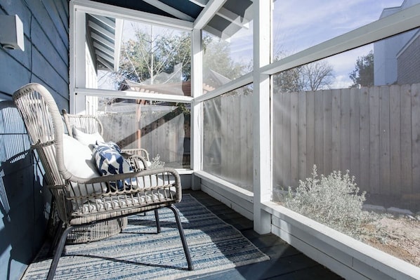A covered patio with a wicker chair, a blue and white patterned rug, and a view of a wooden fence.