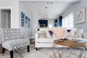 A modern living room with a patterned armchair, a white sofa with decorative pillows, a wooden coffee table, and a kitchen in the background.