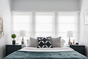 A bedroom with a bed, two bedside tables, a window with blinds, and a wall-mounted artwork.