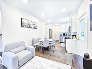 Ein modernes Wohnzimmer mit einer Couch, einem Esstisch, Stühlen, einer Küchenzeile inklusive Geräte und einem gerahmten Bild an der Wand.