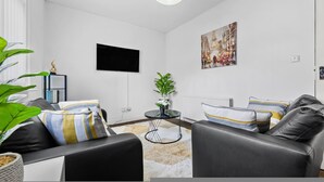 A modern living room with a black leather sofa, a round coffee table, and a flat-screen TV mounted on the wall. - Comfort Studio Leeds Ideal for Work Study Street Parking (Leeds)