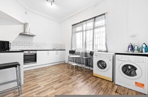 A kitchen with a microwave, oven, and washing machine, a dining area with chairs, and a window with sheer curtains. - Modern Ensuite Studio Leeds Free Parking (Leeds)