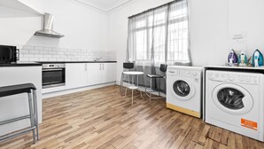 Uma cozinha moderna com micro-ondas, forno e máquina de lavar, piso de madeira, armários brancos e uma janela com cortinas leves.