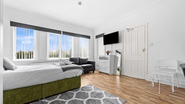 Quarto moderno com cama, escrivaninha, cadeira, televisão e janelas grandes.