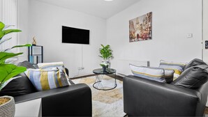 Uma sala de estar moderna com um sofá de couro preto, uma pequena mesa redonda com uma planta, uma televisão de tela plana na parede e um quadro.