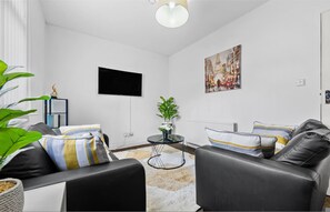 A modern living room with a black leather sofa, a small round table with a plant, a flat-screen TV mounted on the wall, and a painting. - Leeds City Studio Stay Ensuite Kitchenette Street Parking (Leeds)