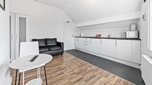 Una cocina moderna con armarios blancos, encimera negra y microondas. Hay un sofá de cuero negro y una mesa blanca con un control remoto.