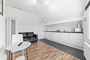 Eine moderne Küche mit weißen Schränken, einer schwarzen Arbeitsplatte und einer Mikrowelle. Ein schwarzes Ledersofa und ein weißer Tisch mit einer Fernbedienung sind ebenfalls vorhanden.