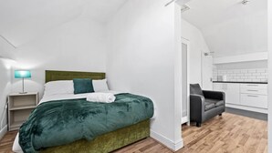 Un dormitorio moderno con una cama tapizada en verde, una lámpara de noche y vistas hacia una zona de cocina.