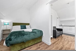Ein modernes Schlafzimmer mit einem mit grünem Stoff bezogenen Bett, einer Nachttischlampe und Blick in einen Küchenbereich.
