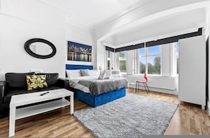 A modern bedroom with a bed, a white sofa, a coffee table, a red chair, a large window, and a cityscape painting. - Cosy Leeds Studio Ensuite Kitchenette Wifi Street Parking (Leeds)