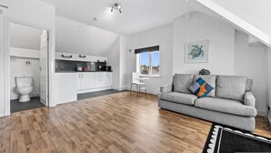 Un espacio habitable moderno con piso de madera, un sofá gris, una zona de comedor blanca y vistas por una ventana.