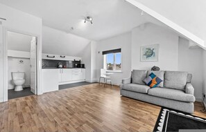 Ein modernes Wohnzimmer mit Holzboden, einer grauen Couch, einem weißen Essbereich und Blick durch ein Fenster.
