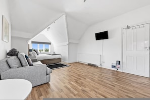 Ein modernes Schlafzimmer im Loft-Stil mit schrägverdeckter Decke, einem großen Bett, einem grauen Sofa, einem Flachbildfernseher und einer weißen Tür.