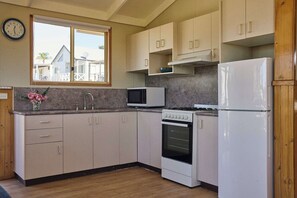 A kitchen with a microwave, oven, and refrigerator, wooden cabinets, a window with a view, and a clock on the wall. - The Mook Mollymook - Morton 53B Cabin (Mollymook)