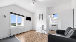 A modern apartment with a wooden floor, a black leather sofa, a TV mounted on the wall, a small table, and a window with a view. - Large Modern Leeds Stay w 8 Private Ensuite Studio (Leeds)