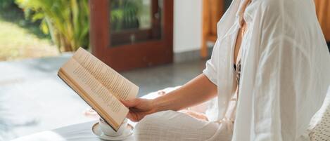 A person sitting on a bed reading a book in a room with a large window and a view of greenery outside.