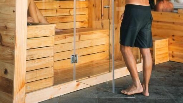 A sauna with wooden walls and a glass door, a bench inside, and a person standing outside. - Co Day Uluwatu  (Pecatu)