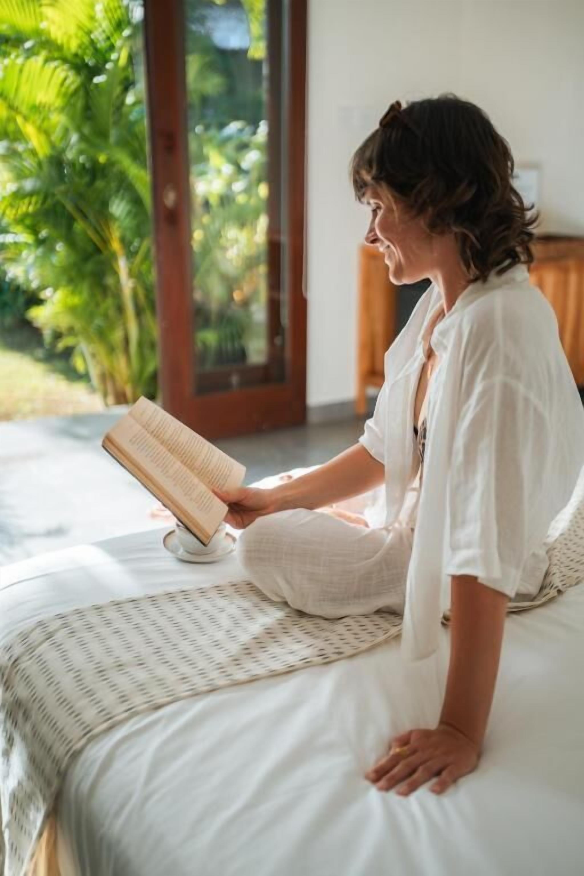 Iemand zit op een bed te lezen in een kamer met een groot raam en uitzicht op groen.