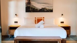 A bedroom with a bed, a bench, two bedside tables, a wall-mounted air conditioner, and a painting. - Co Day Uluwatu  (Pecatu)