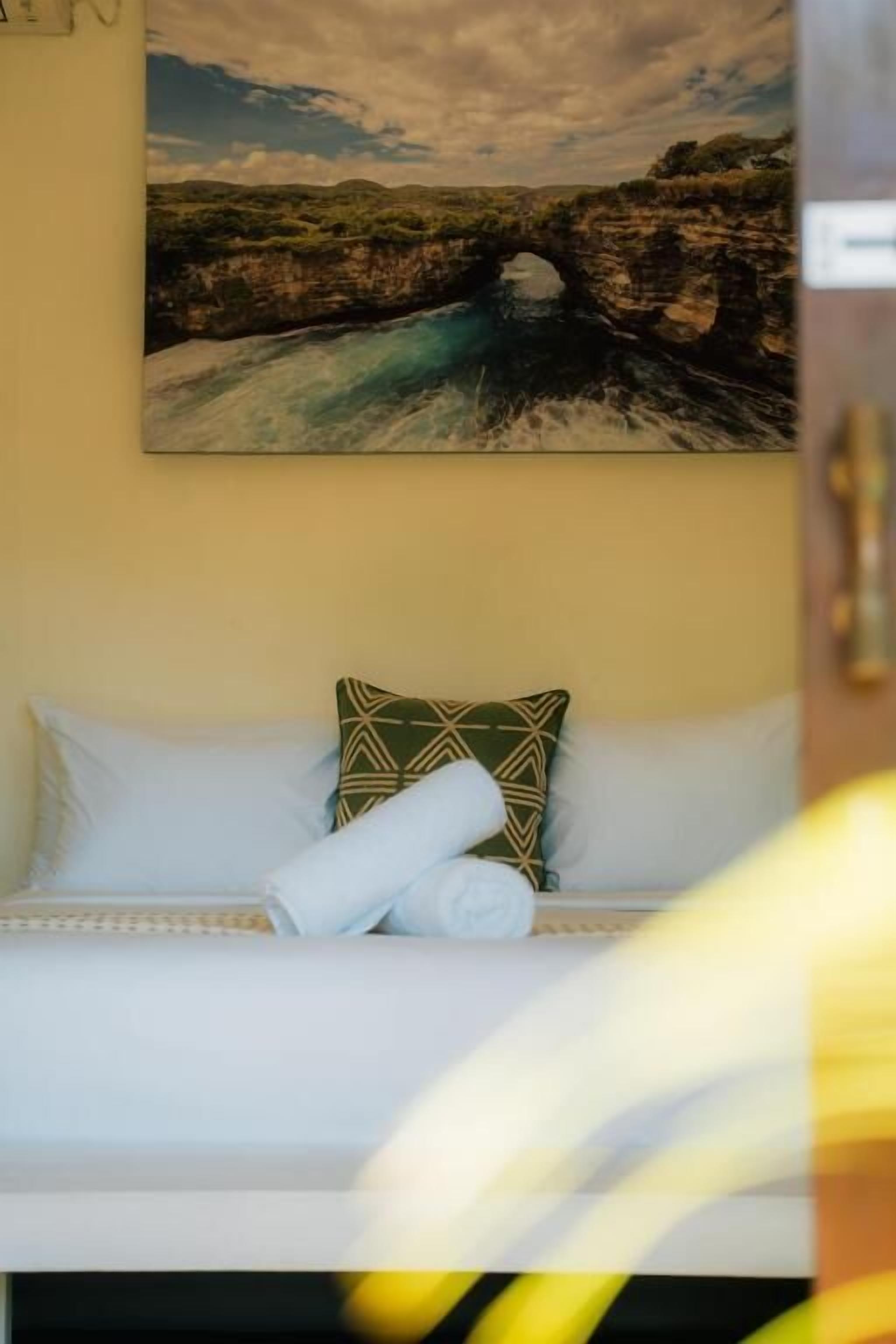 A bed with white bedding, a green patterned pillow, and a rolled white towel.